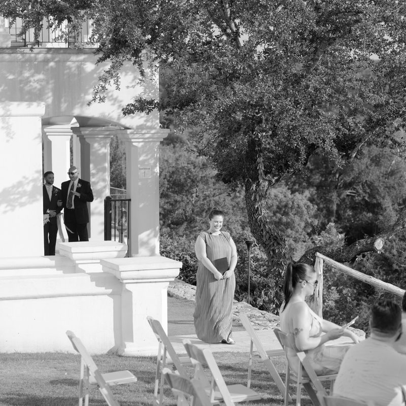 The officiant walks toward the ceremony, script in hand, as groomsmen wait beside white stone columns
