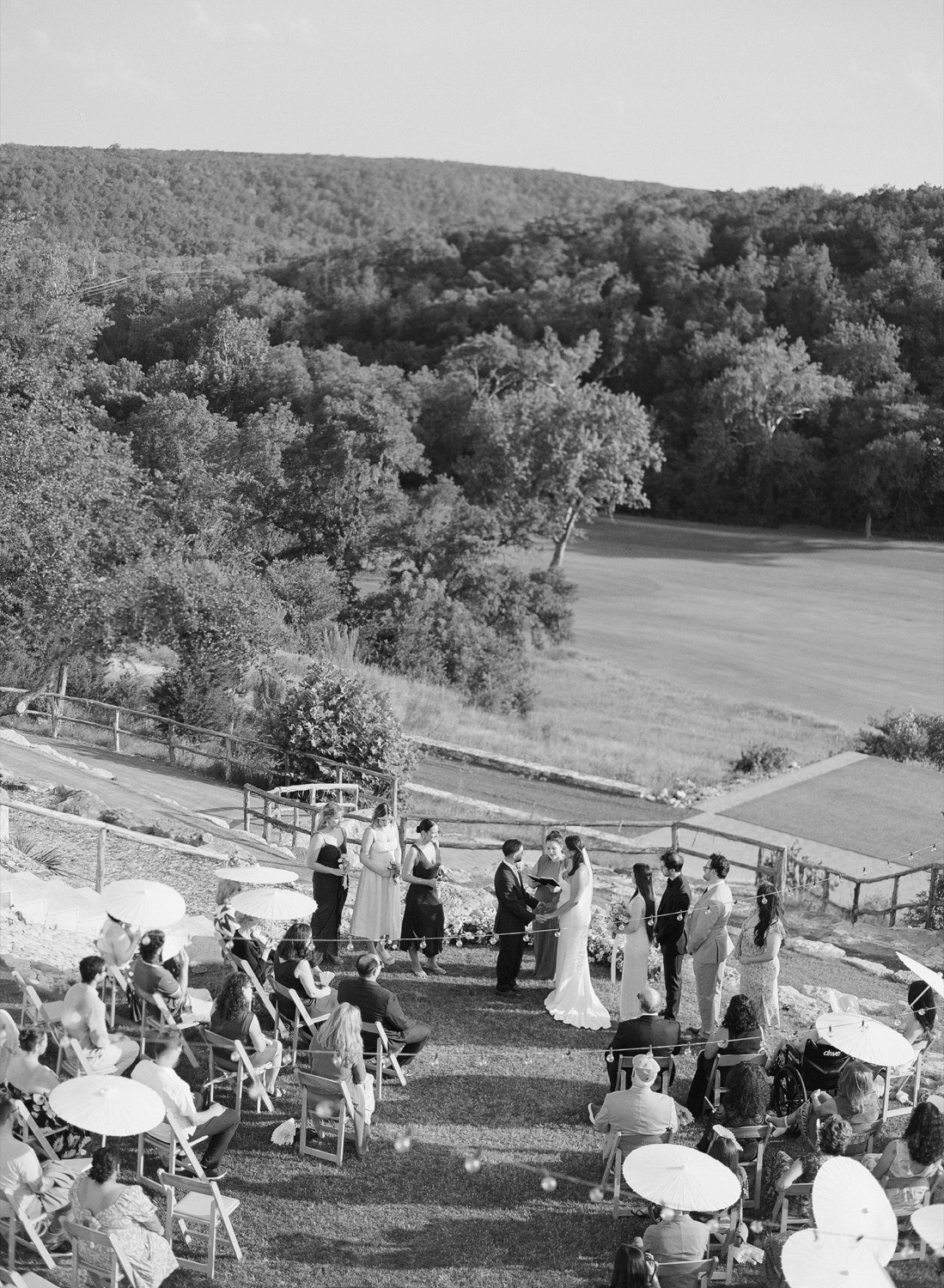 Aerial view of a hillside wedding ceremony overlooking a green valley, guests seated beneath white parasols